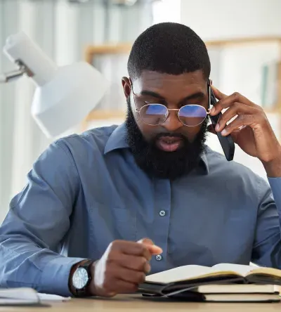 A criminal justice professional in a blue dress shirt and glasses reads a legal book while speaking on the phone, with a scale of justice and a desk lamp in the background.